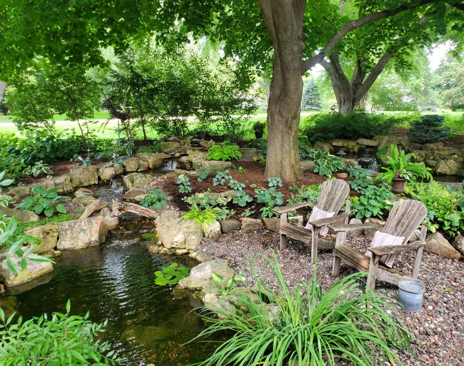 A beautiful water garden with small waterfalls next to a tree with two wooden lawn chairs under it.